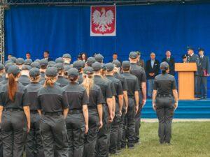 Uczestnicy uroczystości w ciemnych mundurach i czapkach, ustawieni tyłem do widza, stoją na trawie przed sceną. Na scenie, na tle niebieskiej kurtyny z godłem Polski, mężczyzna w mundurze przemawia z mównicy, obok stoją inni oficjele i osoby w strojach cywilnych.