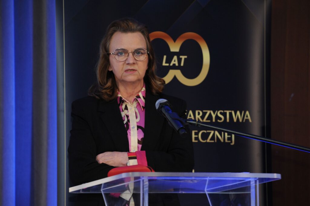 Na zdjęciu, na scenie za mównicą, przed mikrofonem stoi i przemawia Prof. Gertruda Uścińska. W tle na widoczne jest logo wydarzenia.