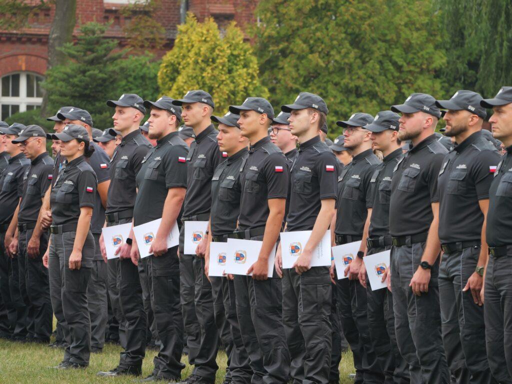 Absolwenci Akademii Wymiaru Sprawiedliwości, funkcjonariusze Służby Więziennej, w mundurach, stojący w rzędach na trawie i trzymający dyplomy podczas uroczystości na świeżym powietrzu.