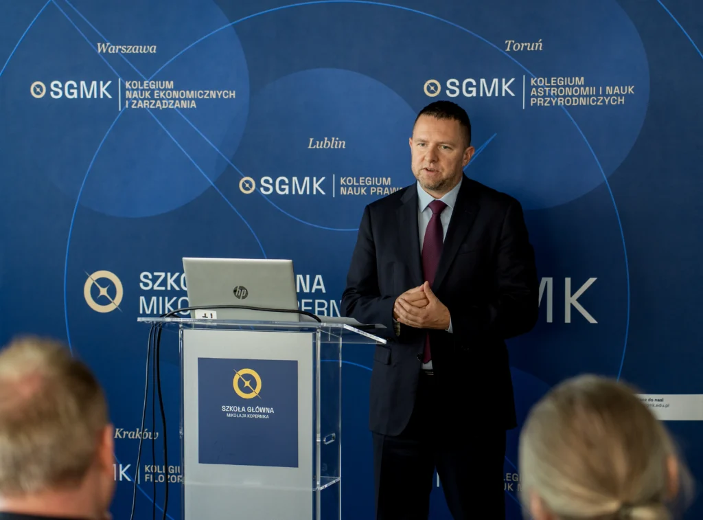 Prelegent w garniturze przemawiający z mównicy, za nim widoczny baner Szkoły Głównej Mikołaja Kopernika. Baner przedstawia listę kolegiów uczelni w różnych miastach, m.in. w Warszawie (Nauk Ekonomicznych i Zarządzania), Toruniu (Astronomii i Nauk Przyrodniczych) oraz Lublinie (Nauk Prawnych), symbolizując jej rozbudowaną strukturę.