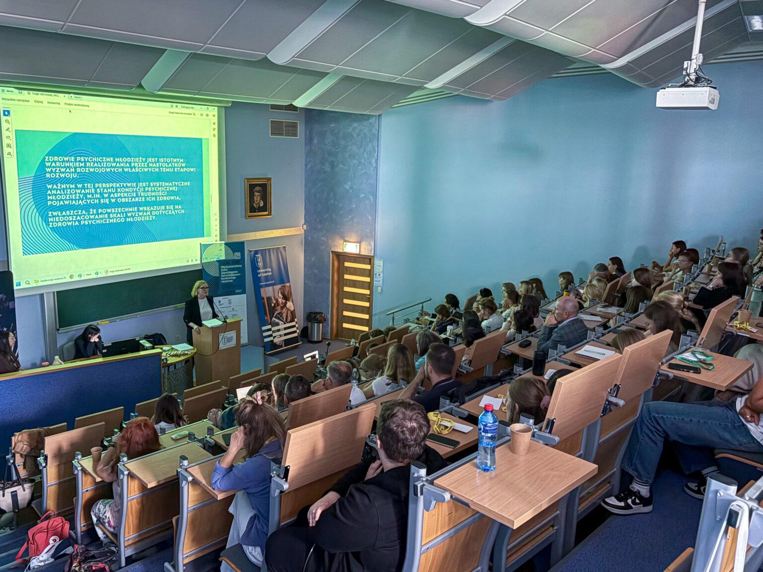 Prezentacja na temat zdrowia psychicznego młodzieży podczas wykładu na Uniwersytecie Gdańskim. W nowoczesnej sali audytoryjnej prelegentka przemawia z podium do słuchaczy siedzących w rzędach, a za nią widoczny jest duży ekran z tekstem dotyczącym treści prelekcji.