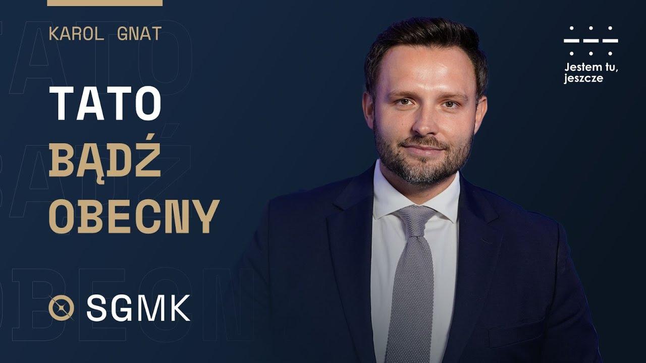 Karol Gnat, uśmiechnięty mężczyzna w garniturze, na ciemnym tle. Po lewej stronie widnieje tytuł "Tato, bądź obecny" oraz logo SGMK. W prawym górnym rogu hasło "Jestem tu, jeszcze".