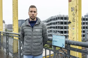 Mężczyzna w szarej kurtce stoi na moście, patrząc prosto w obiektyw z poważnym wyrazem twarzy. Za nim widoczne żółte konstrukcje mostu, miejskie budynki i duży parking wielopoziomowy. Na barierce, obok mężczyzny, znajduje się niebieska tabliczka z napisem "Nie rezygnuj z życia!" oraz numerami telefonów wsparcia i ikoną słuchawki.