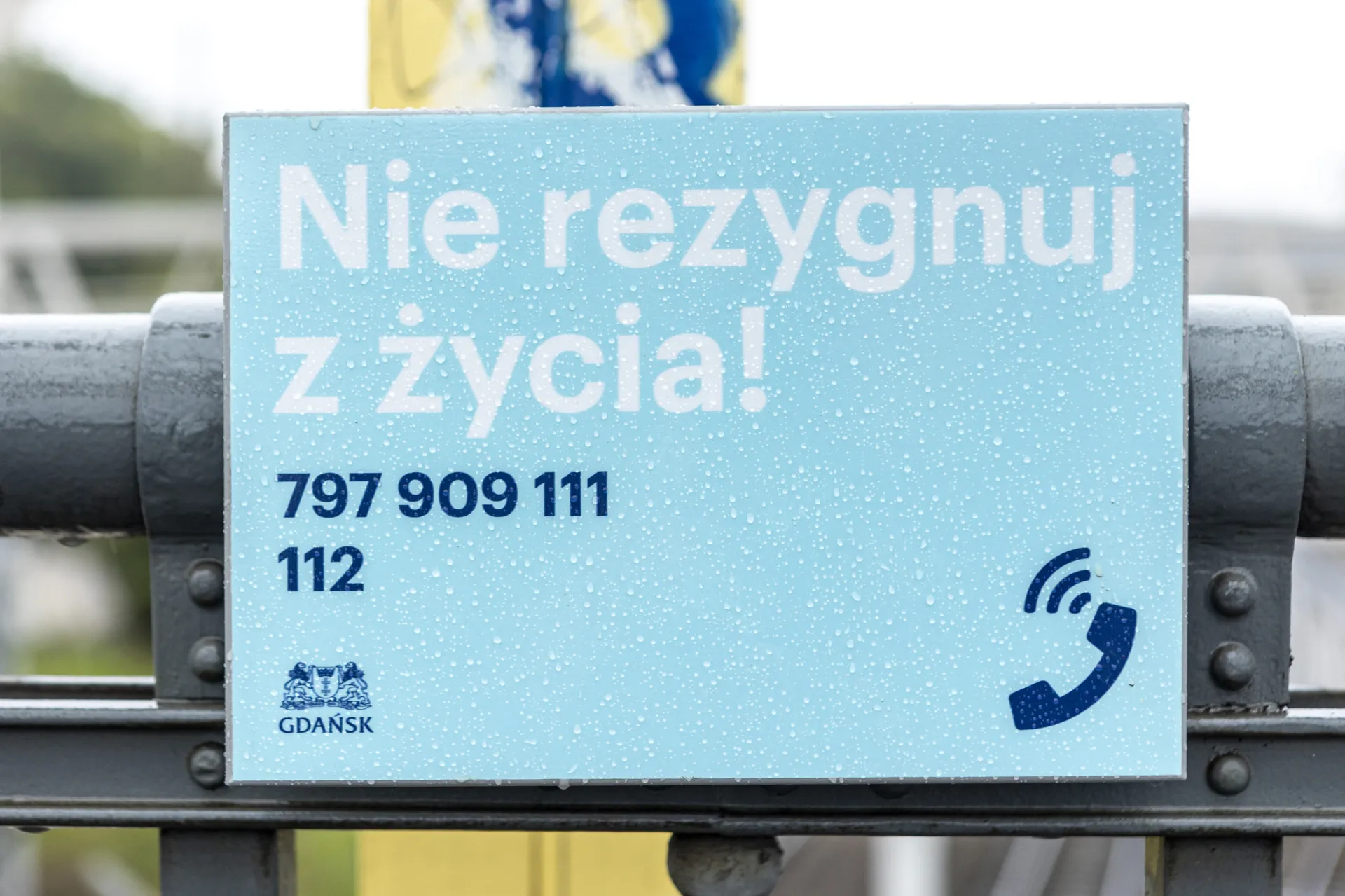 Niebieska tabliczka z tekstem antykryzysowym „Nie rezygnuj z życia!”, numerami wsparcia 797 909 111 i 112, ikoną telefonu oraz logo miasta Gdańsk. Tabliczka jest pokryta kroplami deszczu.