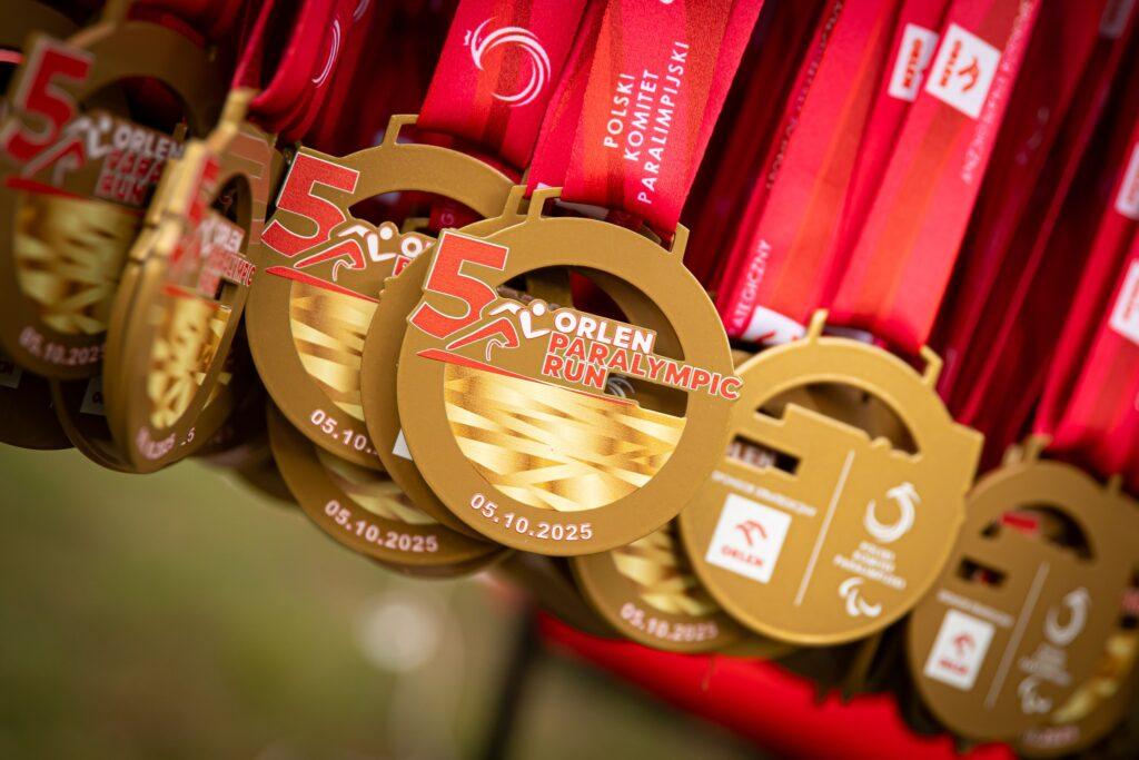 Złote medale pamiątkowe z czerwonymi wstążkami dla Orlen Paralympic Run. Na nich widnieje duża cyfra 5, data 05.10.2025, stylizowana postać biegacza oraz logo Polskiego Komitetu Paraolimpijskiego.