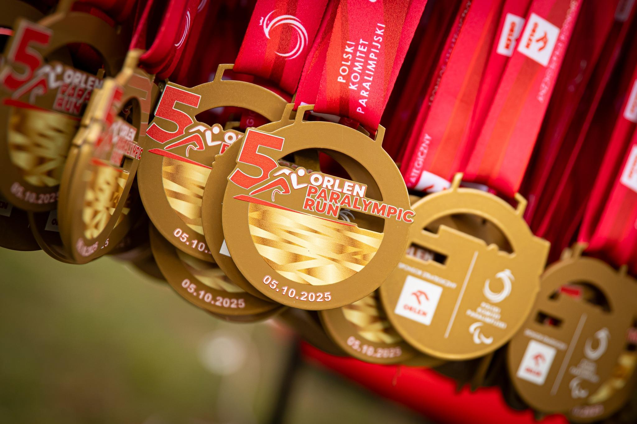 Złote medale pamiątkowe z czerwonymi wstążkami dla Orlen Paralympic Run. Na nich widnieje duża cyfra 5, data 05.10.2025, stylizowana postać biegacza oraz logo Polskiego Komitetu Paraolimpijskiego.
