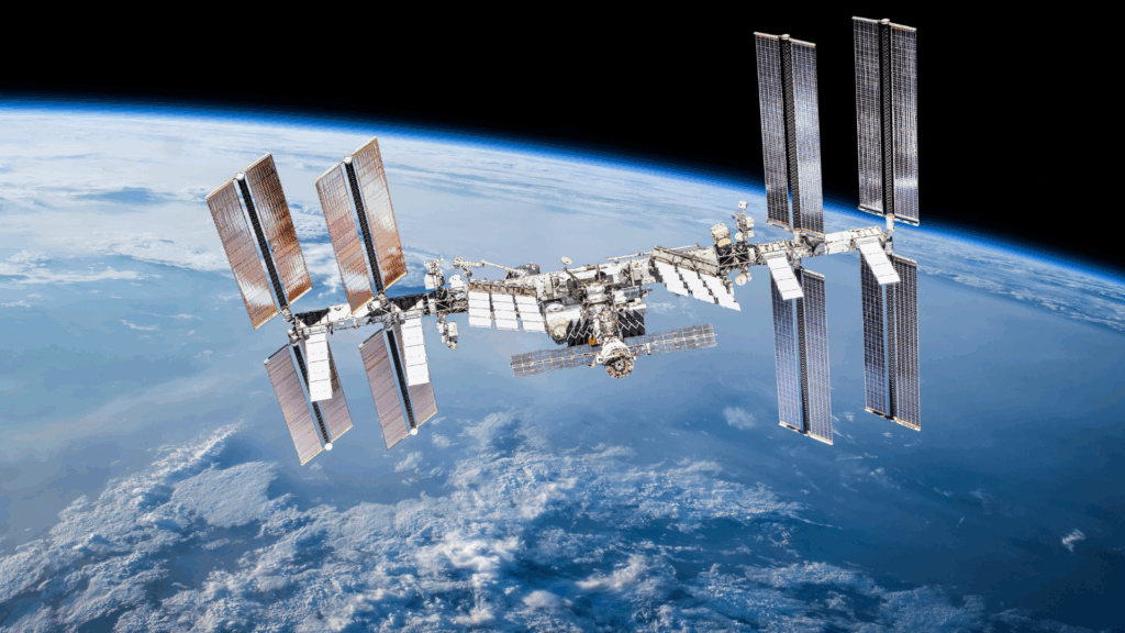 Międzynarodowa Stacja Kosmiczna (ISS) rozciągająca się na tle Ziemi. Widoczne są jej moduły, struktury kratownicowe oraz liczne, rozłożone panele słoneczne. Poniżej rozciąga się błękitna planeta z warstwą atmosfery i białymi chmurami, a powyżej ciemna przestrzeń kosmiczna.