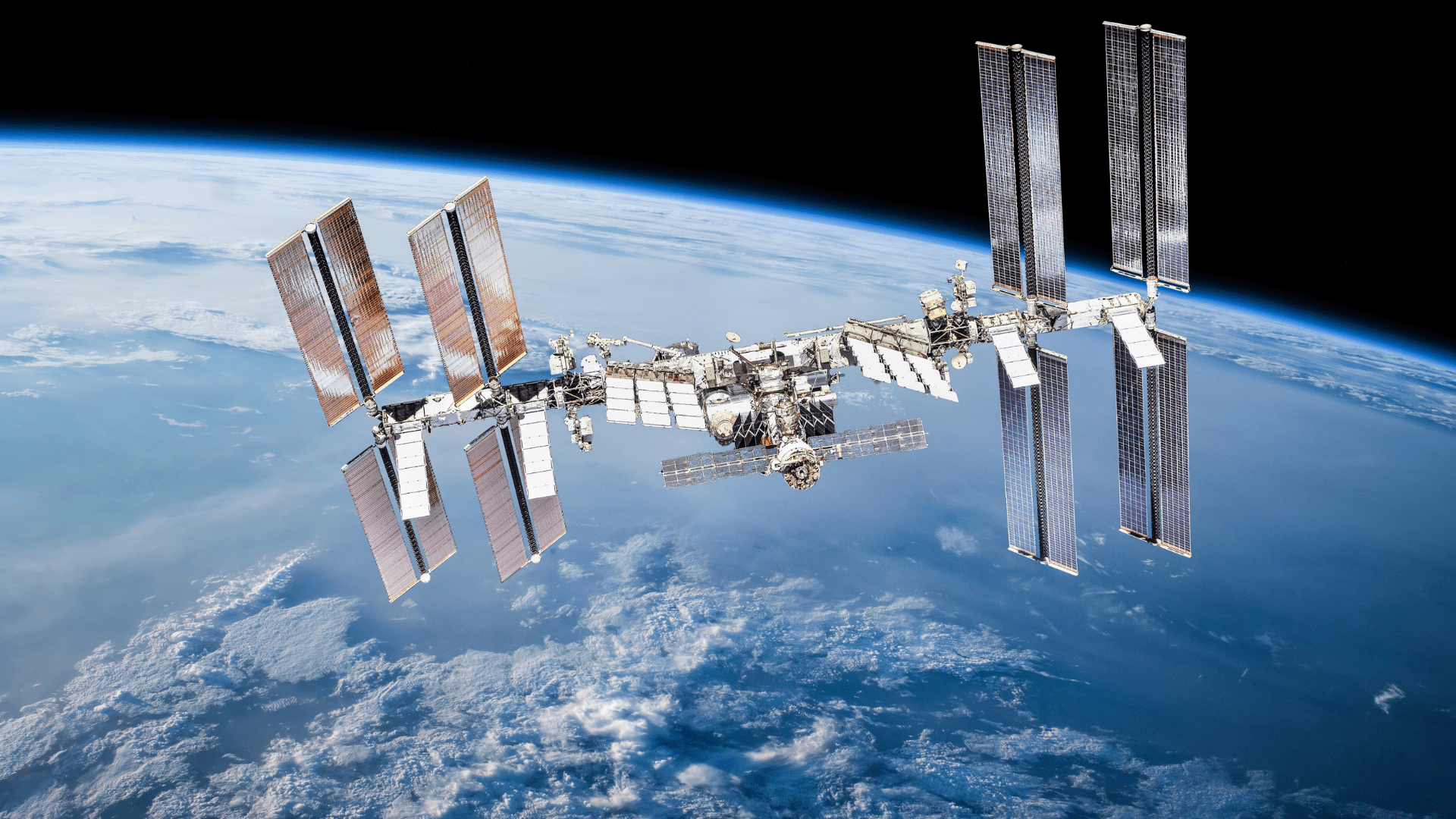 Międzynarodowa Stacja Kosmiczna (ISS) rozciągająca się na tle Ziemi. Widoczne są jej moduły, struktury kratownicowe oraz liczne, rozłożone panele słoneczne. Poniżej rozciąga się błękitna planeta z warstwą atmosfery i białymi chmurami, a powyżej ciemna przestrzeń kosmiczna.