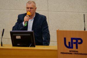 Mężczyzna w ciemnej marynarce przemawia do mikrofonu podczas prezentacji. Na drewnianej mównicy widnieje niebieskie logo UPP oraz napis "WYDZIAŁ PEDAGOGIKI I PSYCHOLOGII".