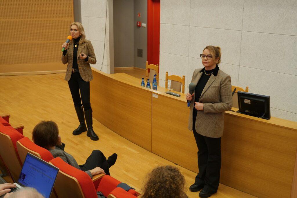 Dwie kobiety z mikrofonami prowadzą prezentację lub panel dyskusyjny w sali wykładowej.