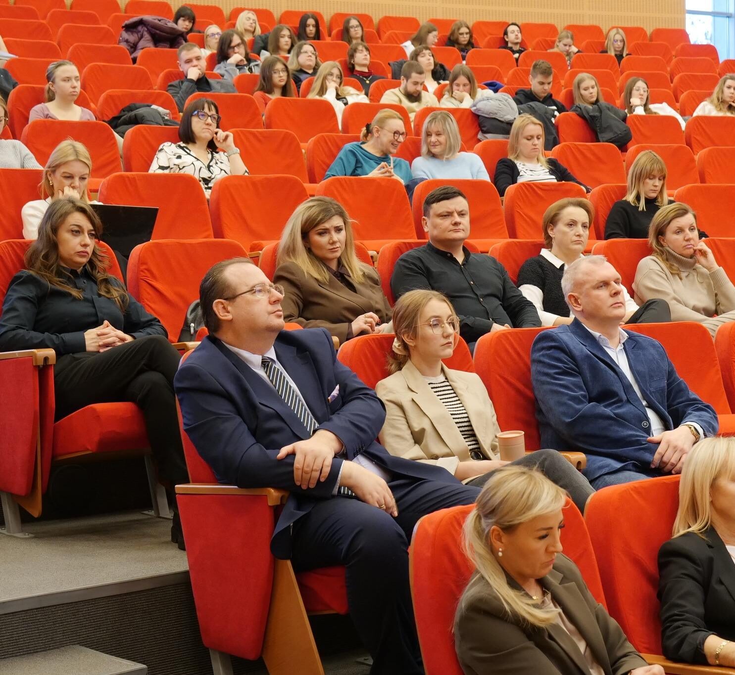 Różnorodna grupa uczestników, w tym kobiety i mężczyźni, siedzi w rzędach pomarańczowych foteli w audytorium, słuchając prezentacji.