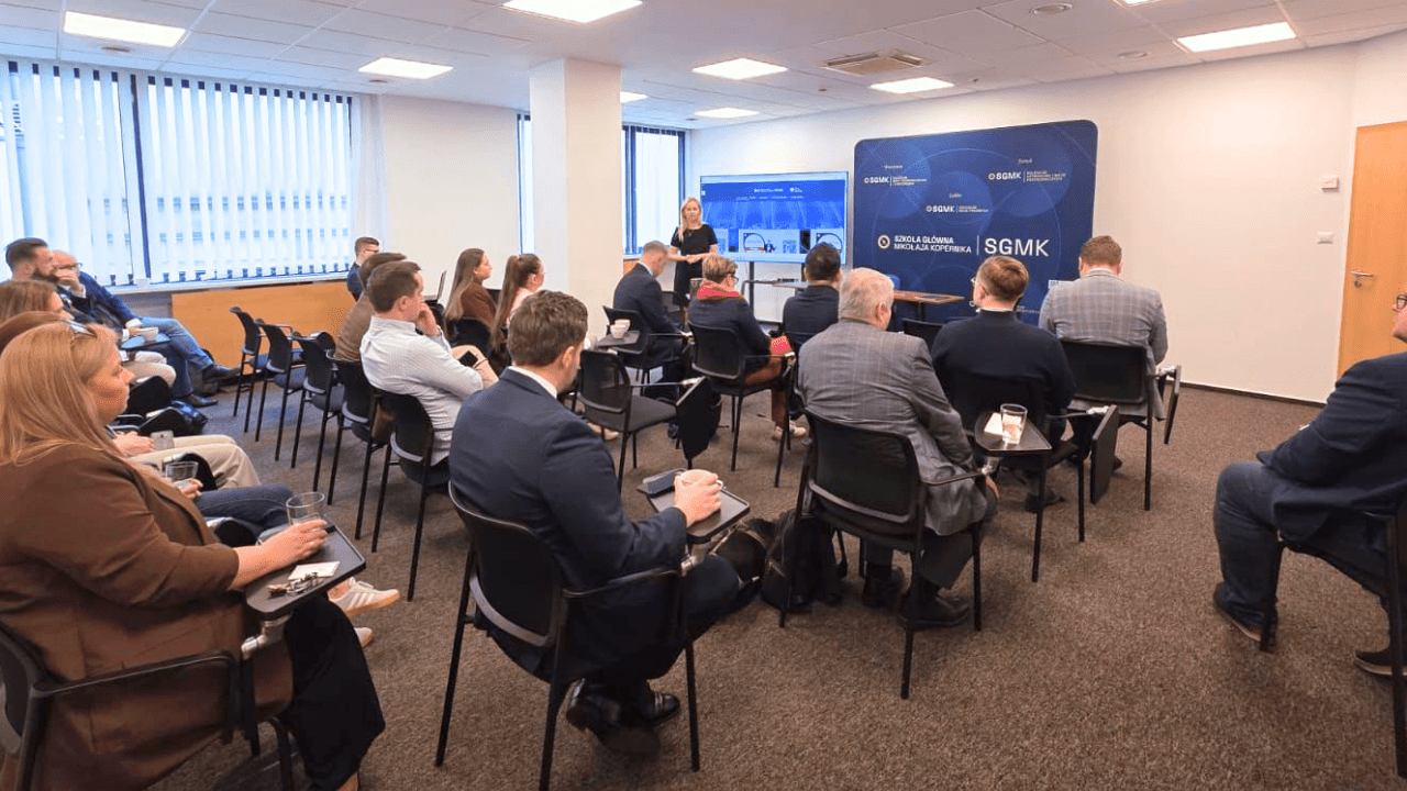Prelegent stoi przed grupą siedzącą na krzesłach w sali szkoleniowej i prowadzi prezentację