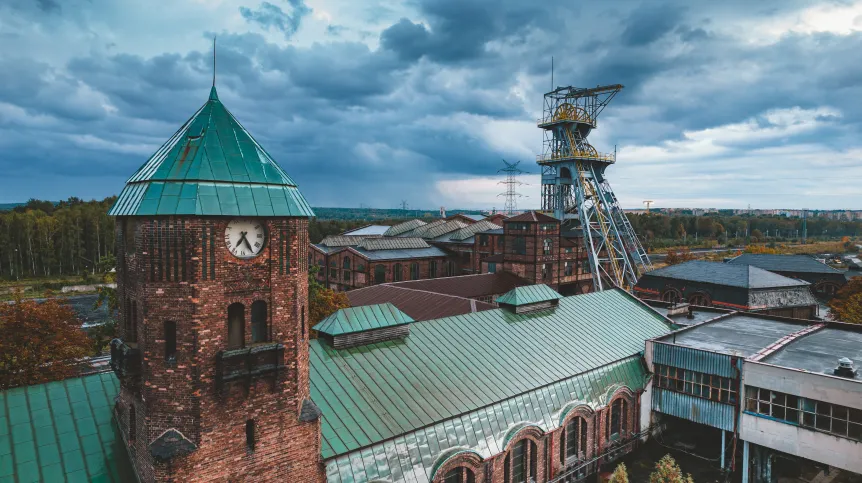 Zabytkowy kompleks górniczy z ceglanymi budynkami i zielonymi dachami. Dominują wieża z zegarem po lewej i stalowy szyb kopalniany po prawej. Scena pod zachmurzonym niebem, z lasem i liniami energetycznymi w tle.