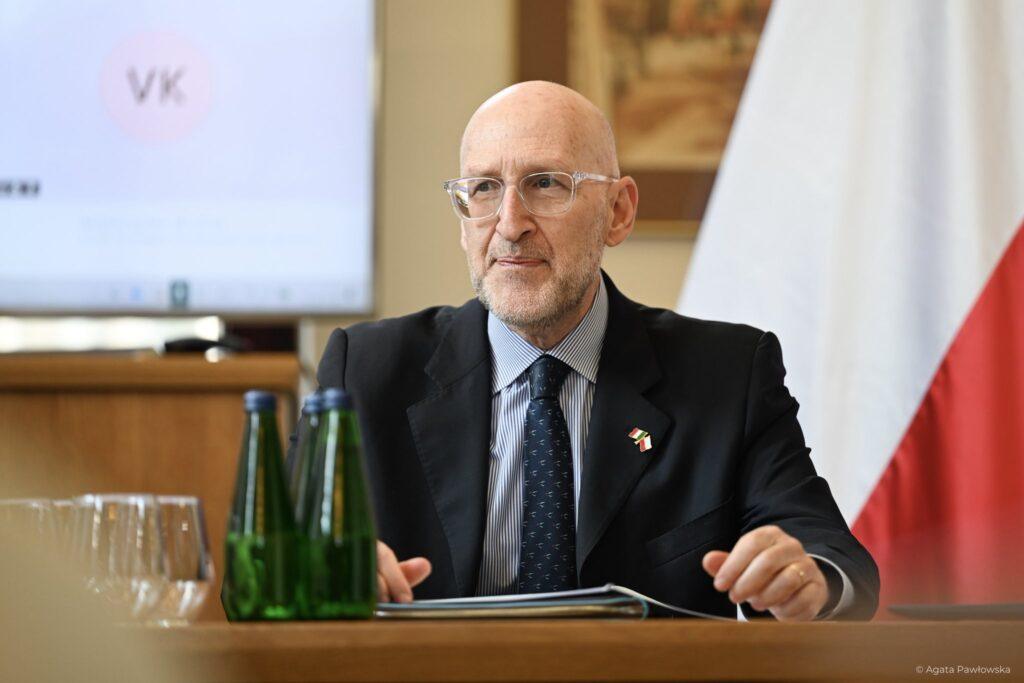 Ambasador Włoch, Łysy, brodaty mężczyzna w okularach i ciemnym garniturze z przypinką flag Polski i Włoch, siedzi przy stole konferencyjnym, patrząc z uwagą. W tle widoczna flaga Polski oraz ekran na których widać połączonych online doktorantów z Włoch.