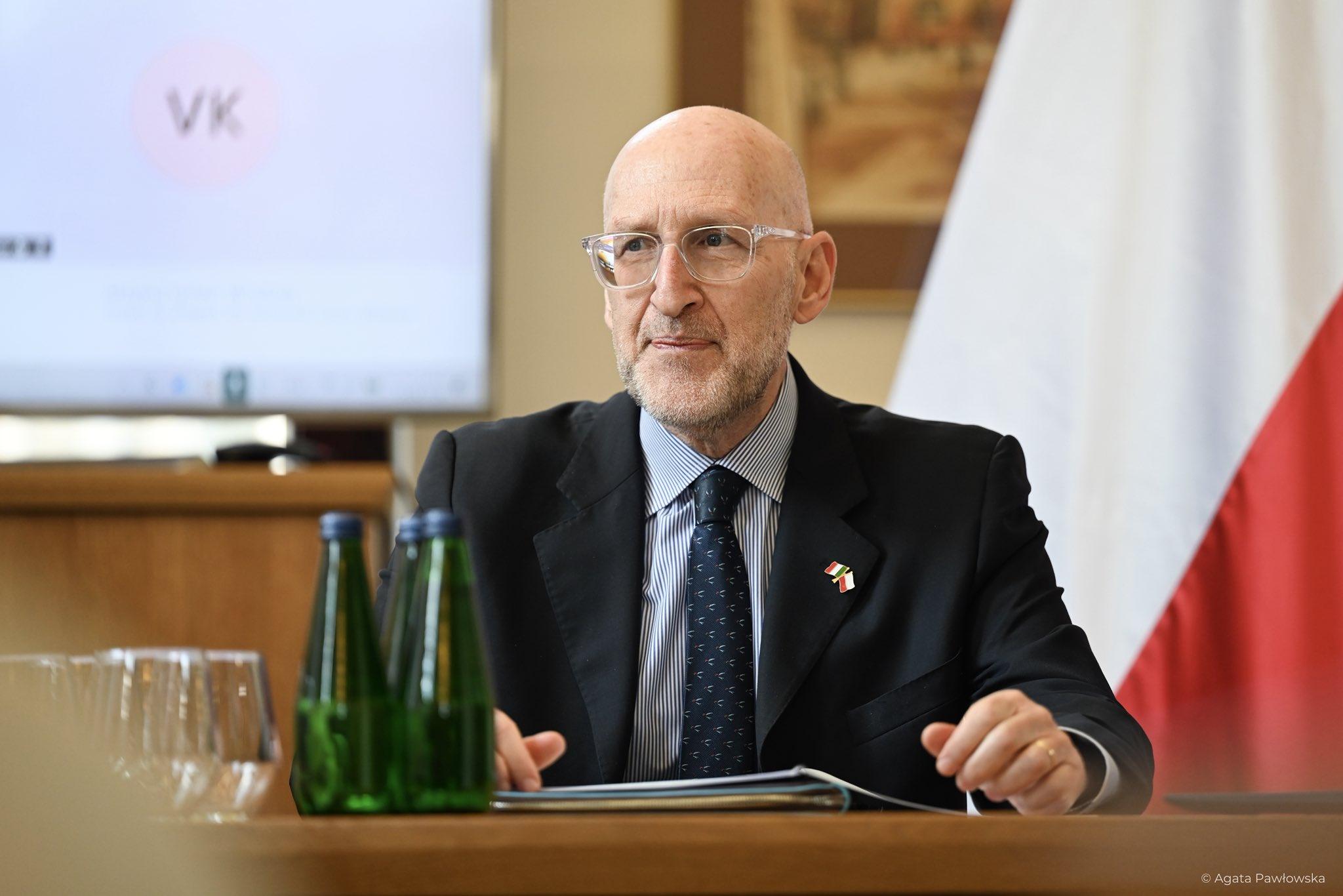 Ambasador Włoch, Łysy, brodaty mężczyzna w okularach i ciemnym garniturze z przypinką flag Polski i Włoch, siedzi przy stole konferencyjnym, patrząc z uwagą. W tle widoczna flaga Polski oraz ekran na których widać połączonych online doktorantów z Włoch.