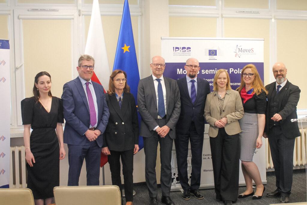 Dziewięć osób w strojach formalnych stoi razem do grupowego zdjęcia. W tle widoczne są flagi Polski i Unii Europejskiej oraz banery promujące seminarium z logotypami Instytutu Pracy i Spraw Socjalnych (IPiSS), Komisji Europejskiej oraz projektu MoveS.