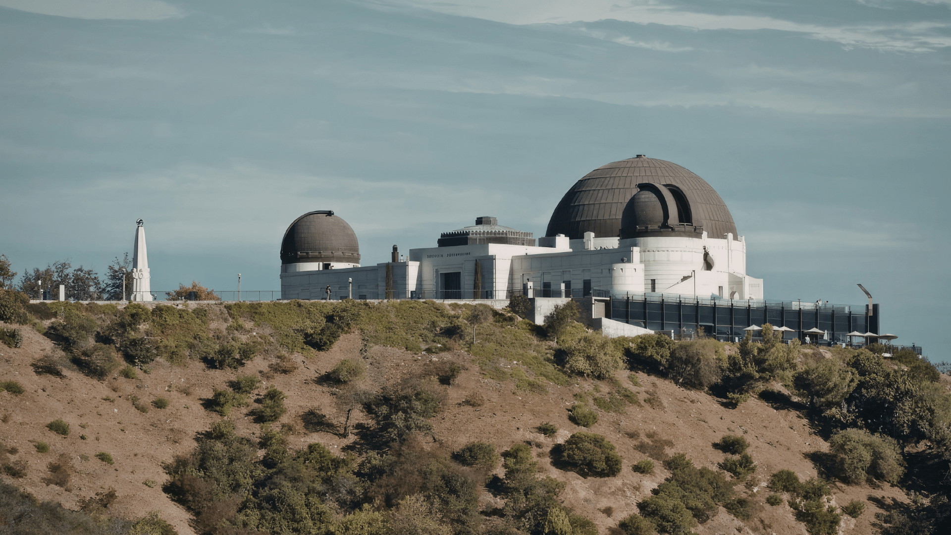 Obserwatorium Griffitha w Los Angeles: biały budynek w stylu Art Deco z trzema kopułami, usytuowany na szczycie porośniętego krzewami wzgórza. Z lewej strony widoczny biały obelisk, w tle częściowo zachmurzone niebo.