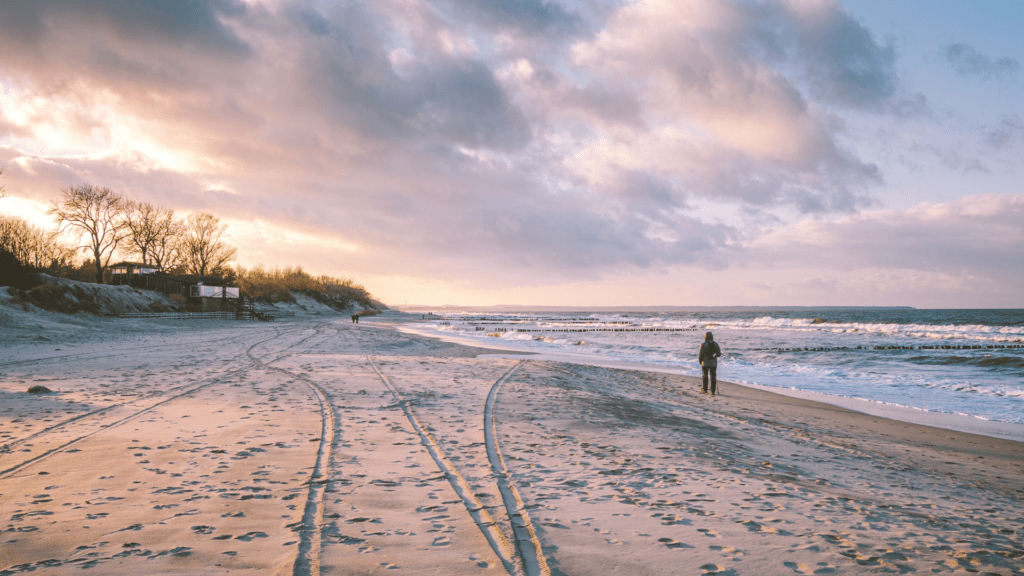 Plaża o zachodzie słońca z osobą spacerującą wzdłuż brzegu morza. W tle fale, drewniane ostrogi i zalesione wydmy z zabudowaniami, pod różowo-pomarańczowym niebem.