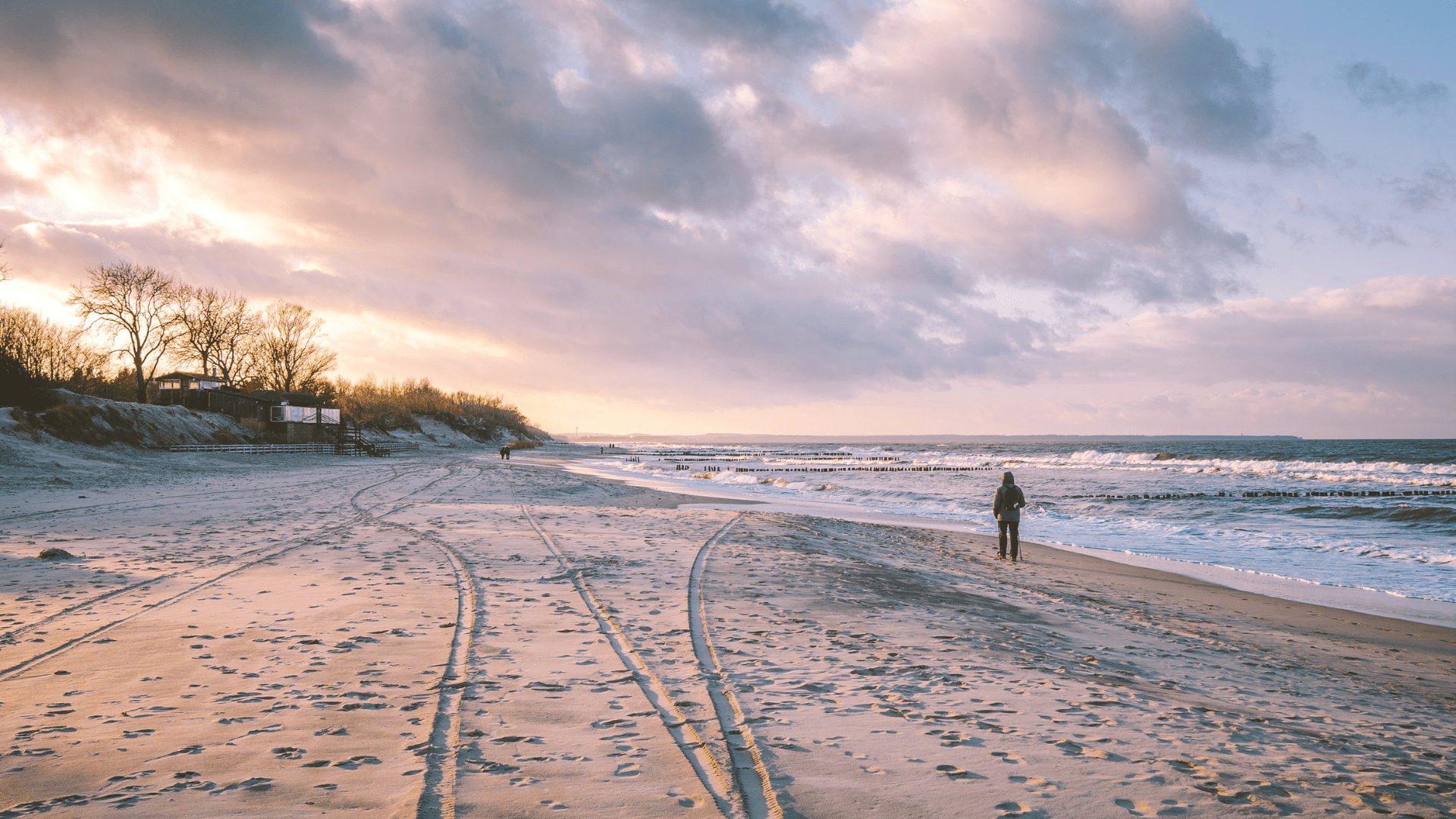 Plaża o zachodzie słońca z osobą spacerującą wzdłuż brzegu morza. W tle fale, drewniane ostrogi i zalesione wydmy z zabudowaniami, pod różowo-pomarańczowym niebem.