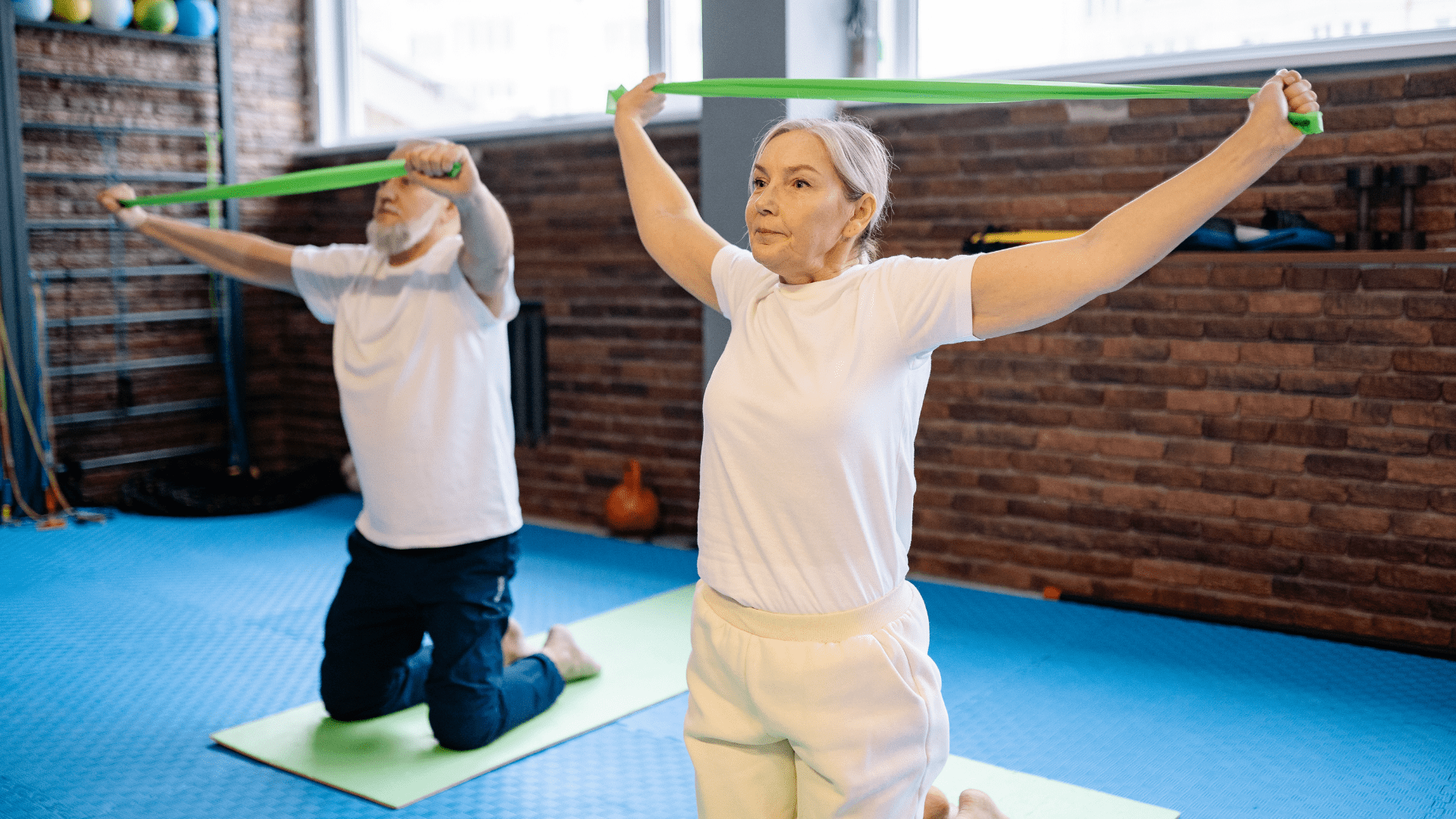 Starsza kobieta i mężczyzna klęczą na matach w sali gimnastycznej, ćwicząc z zielonymi gumami oporowymi, rozciągając je z uniesionymi ramionami. Promocja aktywności fizycznej seniorów.