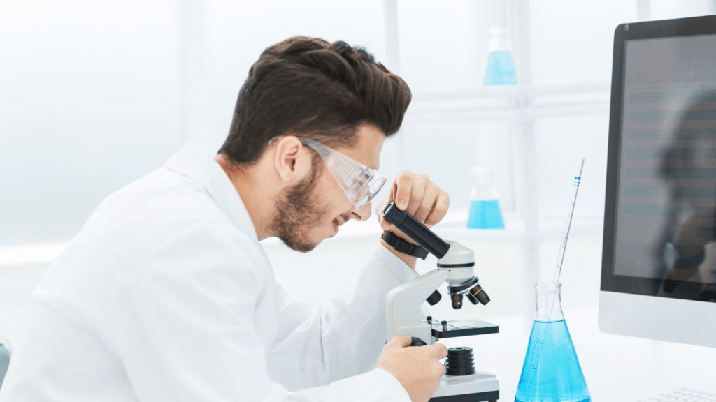 Mężczyzna w białym fartuchu laboratoryjnym i okularach ochronnych obsługuje mikroskop. W tle widoczne kolby z niebieską cieczą i monitor komputera w laboratorium.