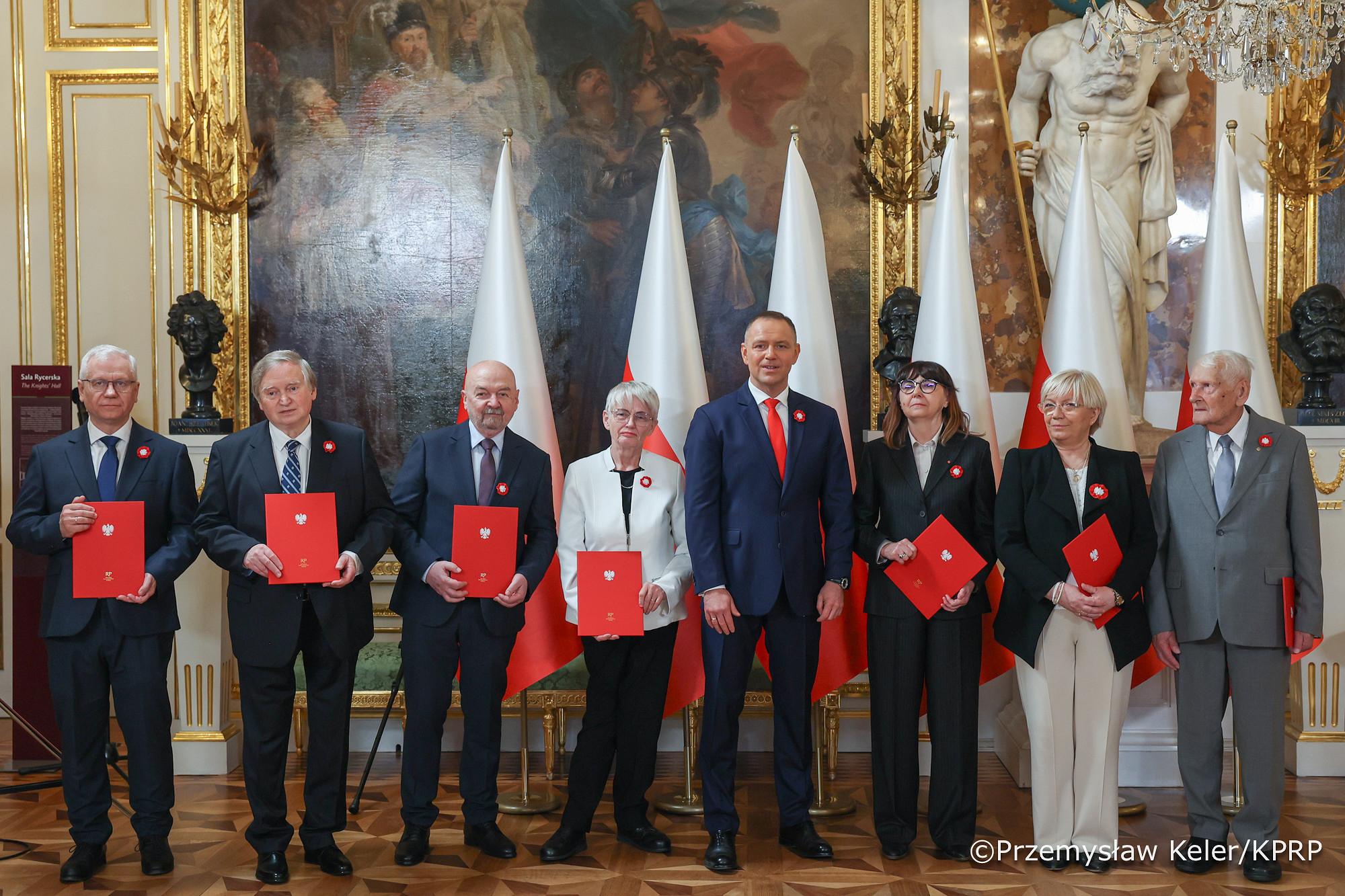 Osiem osób, w tym sześciu mężczyzn i dwie kobiety, stoi w rzędzie w eleganckiej sali. Każdy z nich trzyma czerwoną teczkę z godłem Rzeczypospolitej Polskiej i ma przypiętą biało-czerwoną rozetkę na piersi. W tle widoczne są polskie flagi, obrazy i posągi.