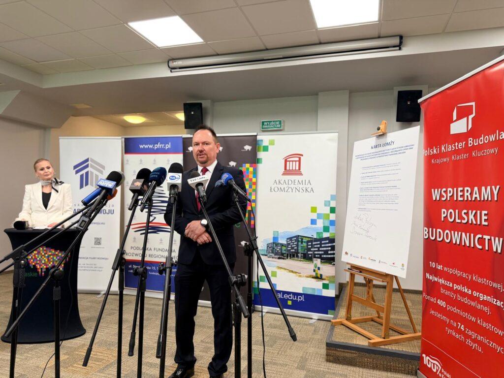 Mężczyzna przemawia podczas konferencji prasowej w Akademii Łomżyńskiej przed mikrofonami mediów.