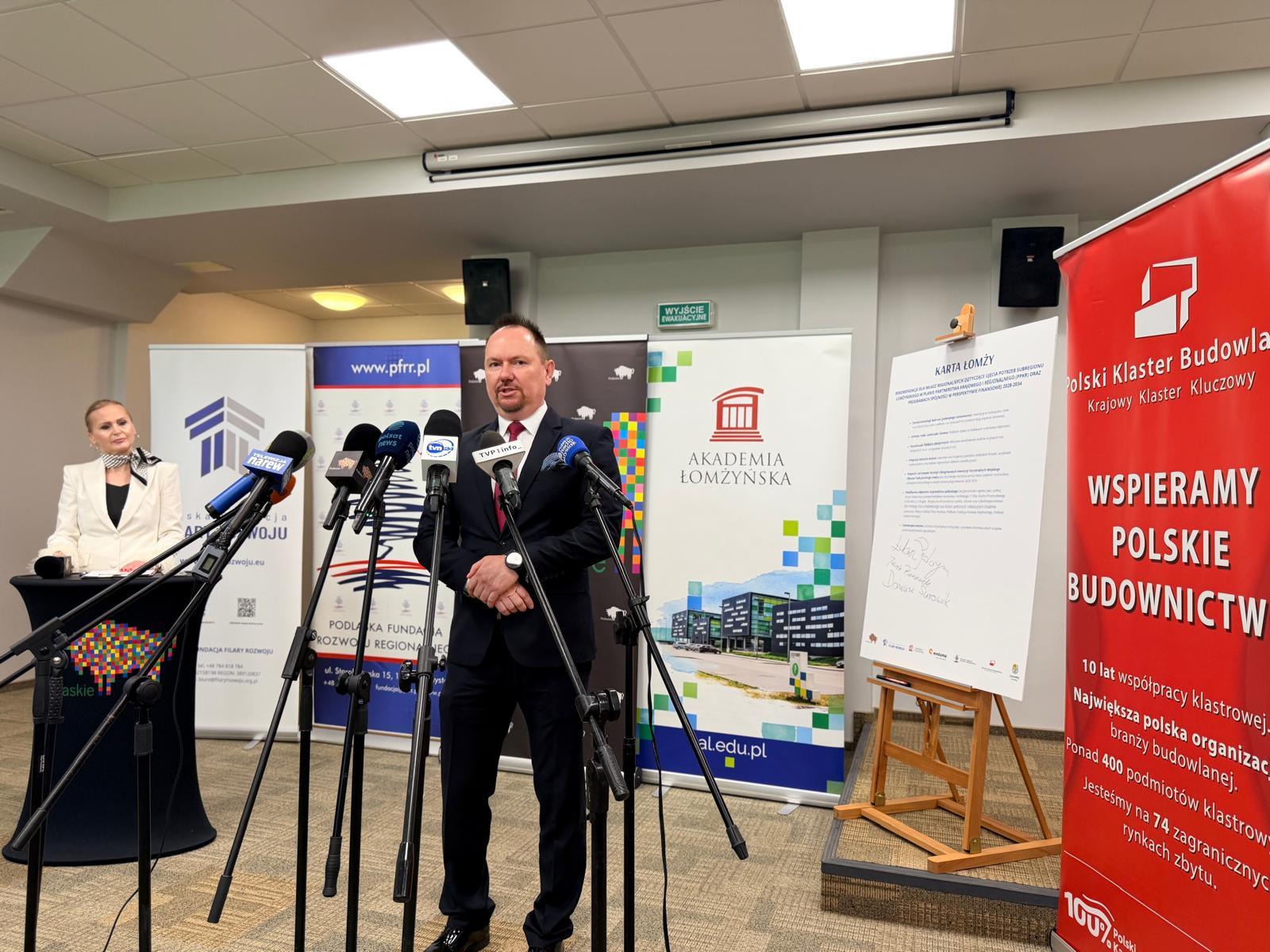 Mężczyzna przemawia podczas konferencji prasowej w Akademii Łomżyńskiej przed mikrofonami mediów.