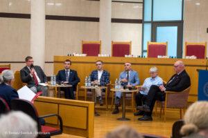 Panel dyskusyjny z udziałem sześciu osób. Jeden z uczestników przemawia do mikrofonu w sali konferencyjnej.