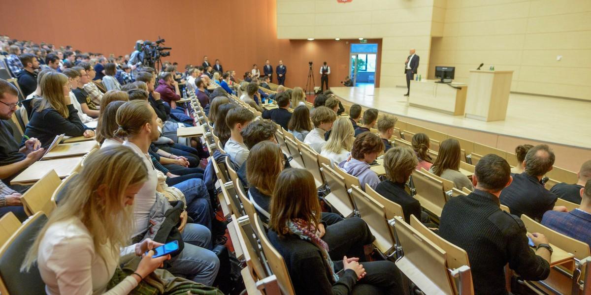 Aula uniwersytecka wypełniona studentami słuchającymi wykładu, z prelegentem na scenie i sprzętem wideo z tyłu sali.