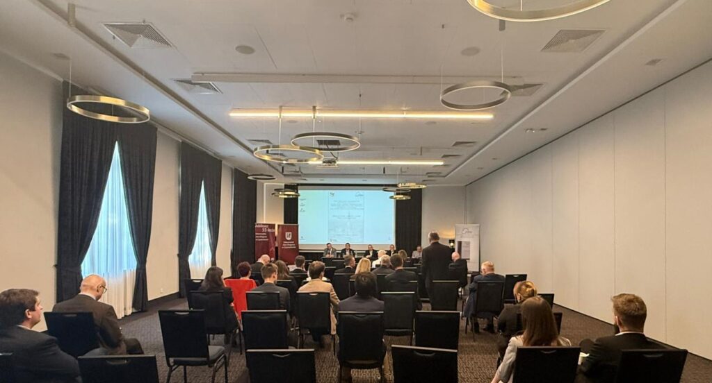 Uczestnicy konferencji siedzący w rzędach przodem do ekranu z prezentacją i panelu prelegentów.