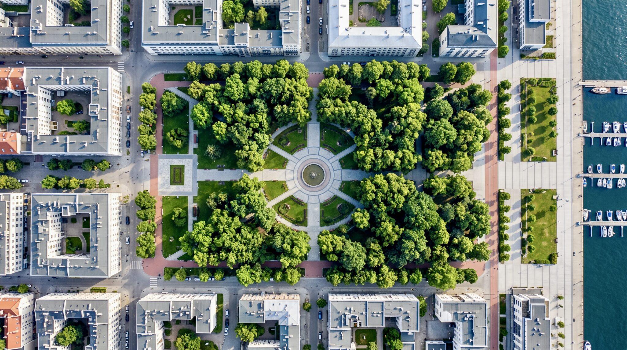 Symetryczny park miejski z lotu ptaka z centralną fontanną, gęstą zielenią, otoczony zabudową i mariną.